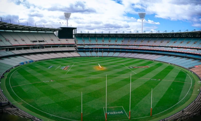 a large round stadium with green grass and white poles with Melbourne Cricket Ground in the background